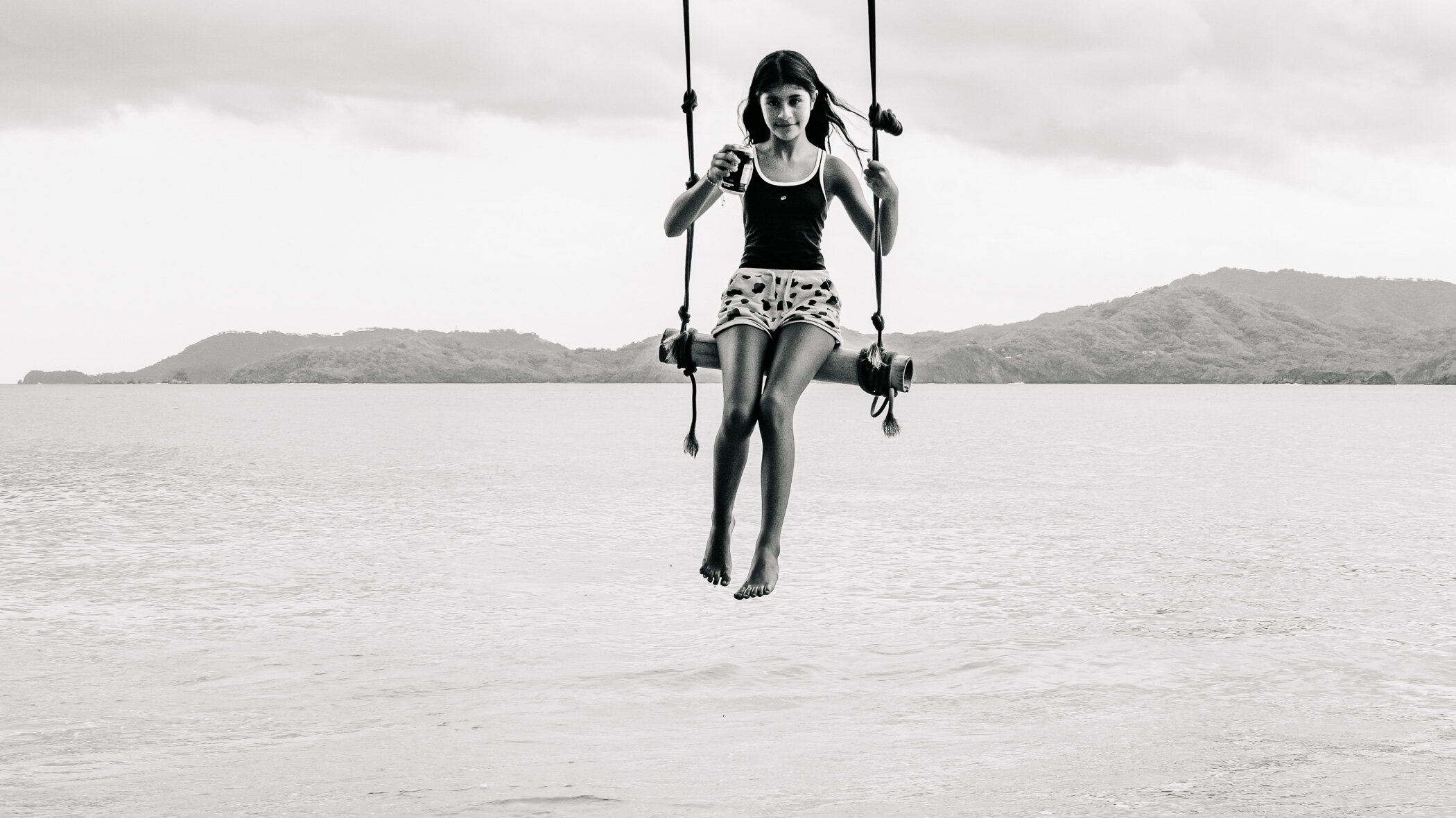 Foto en blanco y negro de una niña en un columpio sobre el agua en la playa